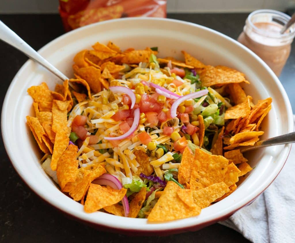 Full salad bowl with two forks sticking out of the bowl on either side, a bag of doritos in the background with a jar of the thousand island dressing