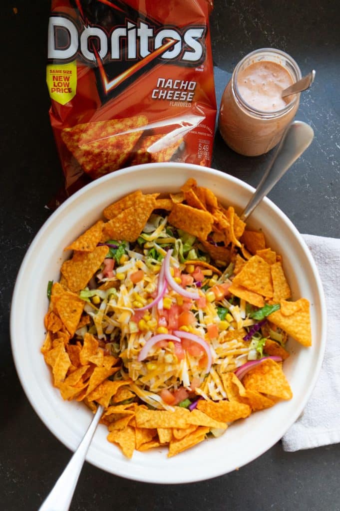 taco salad with dorito chips around the rim of the bowl, bag of doritos in the upper left hand corner and a jar of thousand island dressing in the upper right hand corner