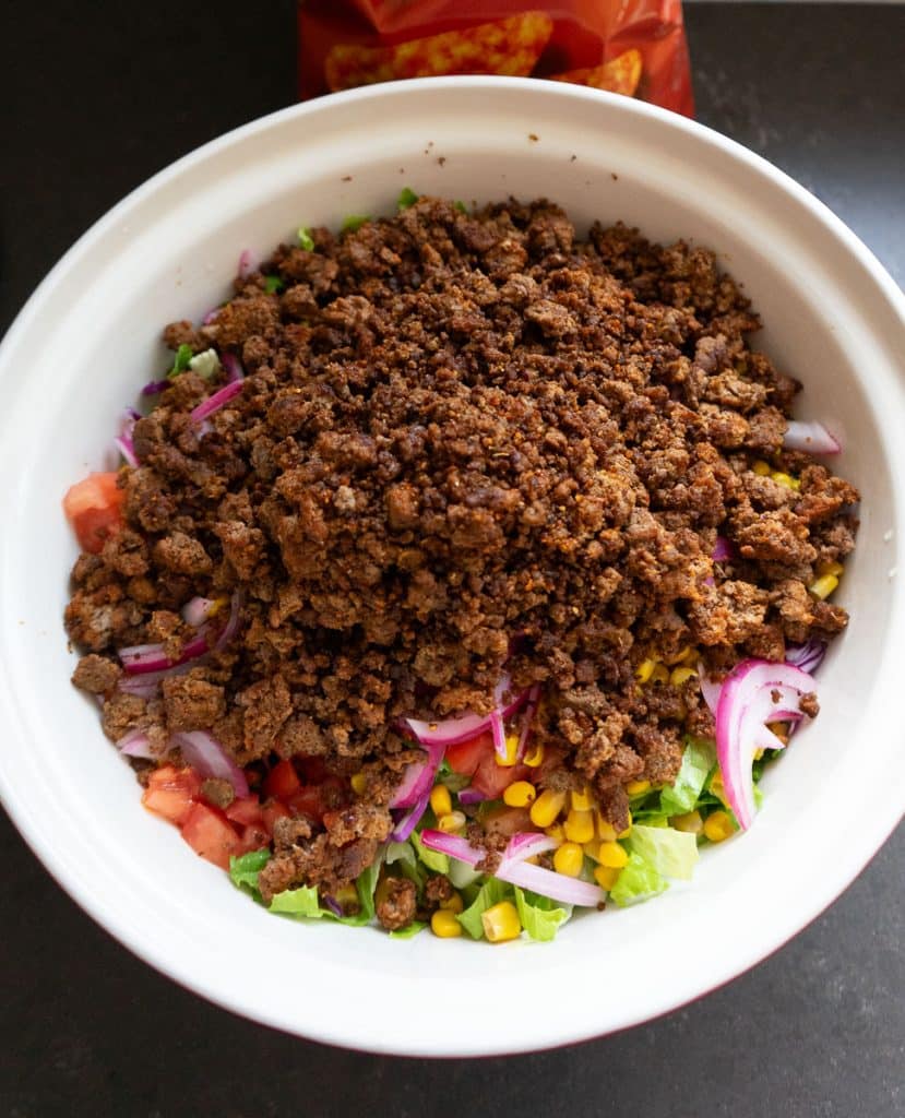 taco meat on top of layered salad ingredients in a large bowl