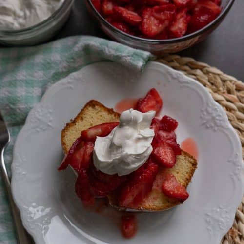 easy classic pound cake with fresh cut strawberries and whipped topping on a white late with bowls of strawberries and whipped cream in the background