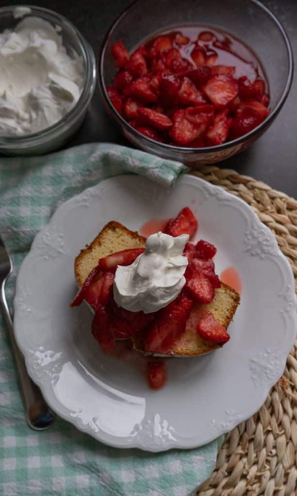 easy classic pound cake with fresh cut strawberries and whipped topping on a white late with bowls of strawberries and whipped cream in the background