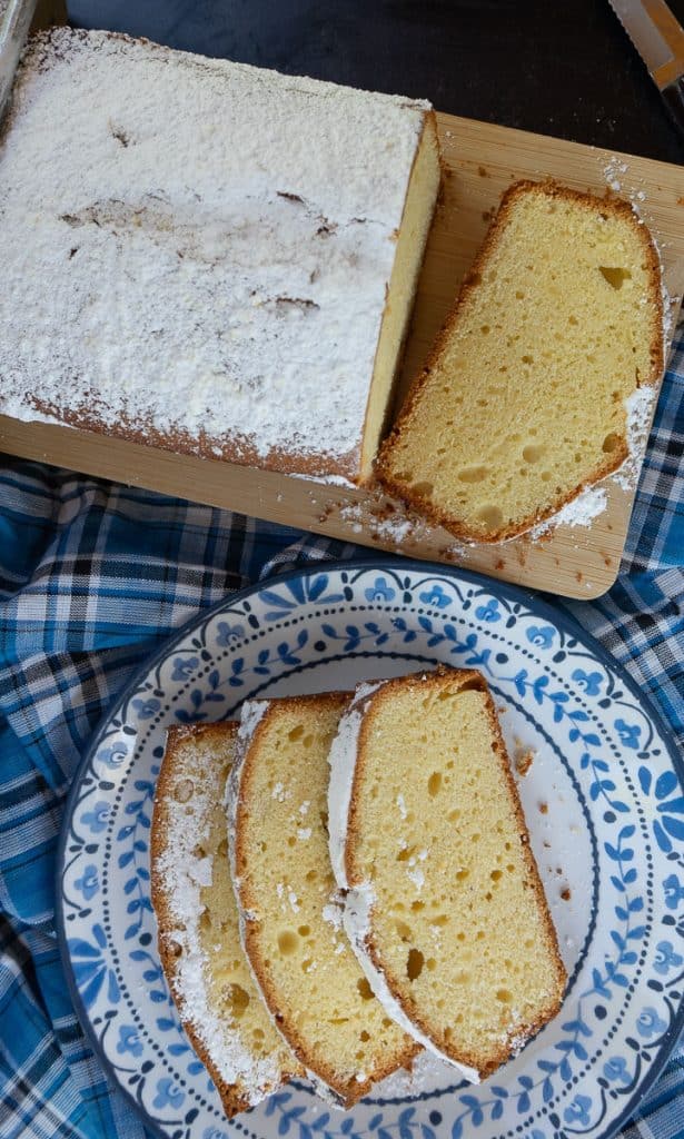 pound cake sliced on a wood cutting board, a blue rimmed plate has three slices of pound cake on it below the wood cutting board in the photo