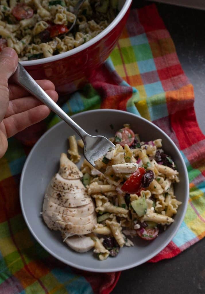 plate of greek pasta salad with sliced chicken on the side next to the large serving bowl of greek pasta salad on a black counter top with a multi colored tea towel under the bowl, a hand is holding a fork full of salad
