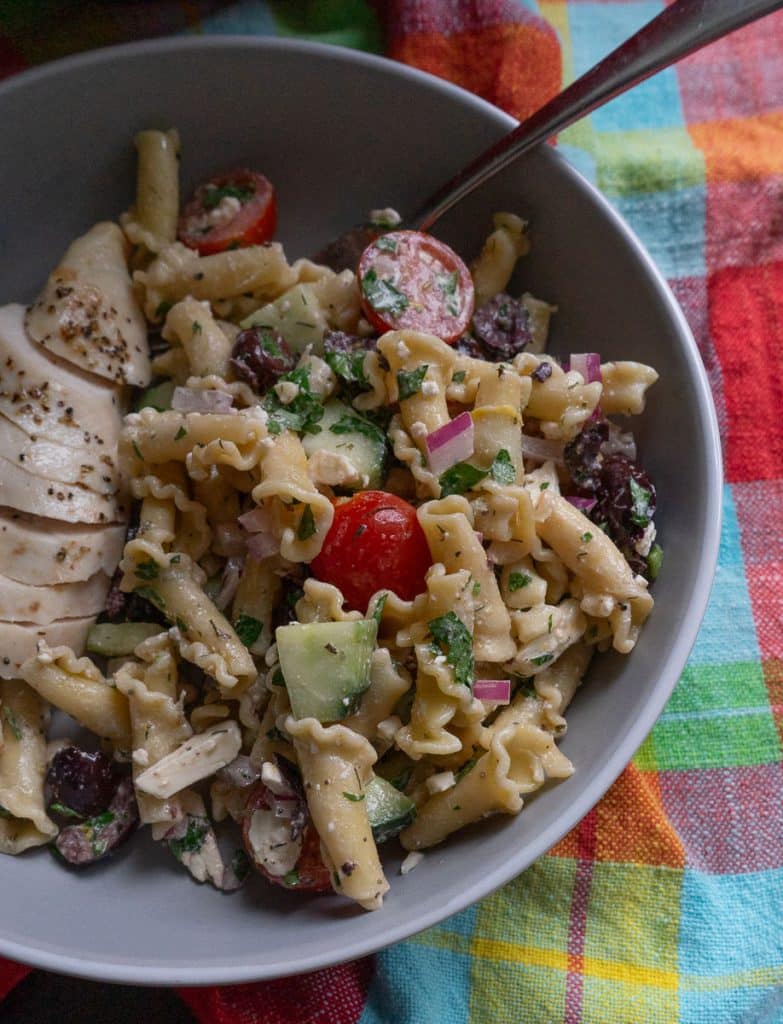 large bowl of greek pasta salad  in a bowl, spoon sticking out of the right side of the bowl, bowl is sitting on a multi colored towel