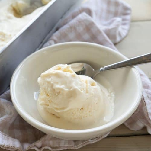 scoop of vanilla ice cream in a white bowl on a picnic table with a tan and white gingham cloth under the bowl
