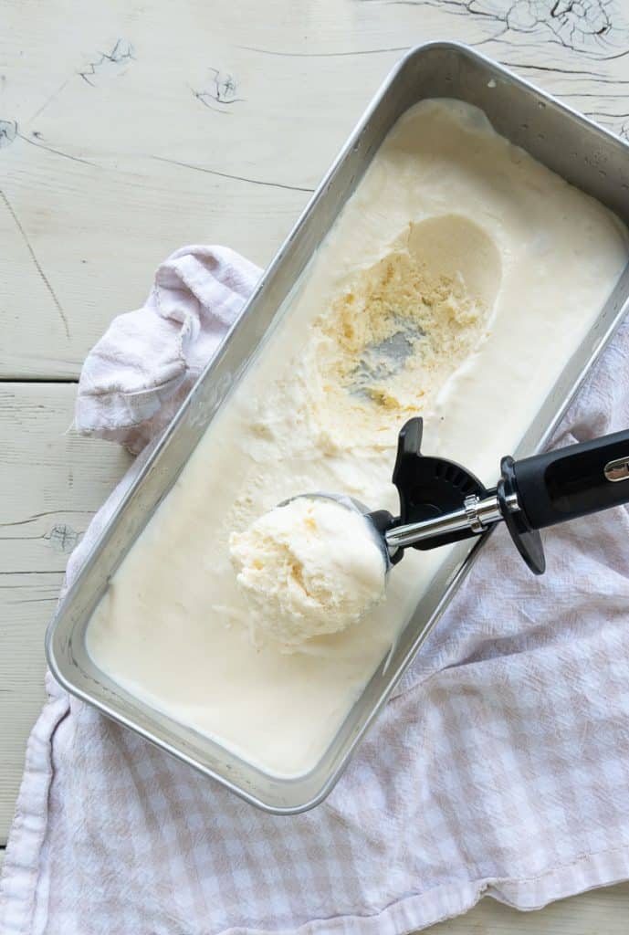 top view of vanilla ice cream in a silver container with a scoop of ice cream taken out and the ice cream scoop laying on top of the ice cream this is sitting on a picnic table 