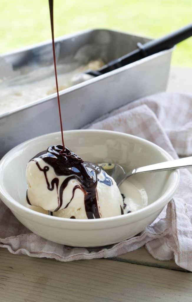 white bowl of vanilla ice cream with chocolate sauce being drizzled from the top silver container of ice cream in the back ground