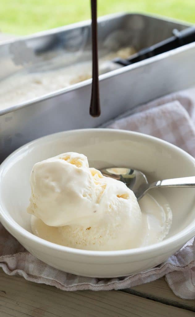 scoop of vanilla ice cream in a white bowl on a picnic table with a tan and white gingham cloth under the bowl with chocolate sauce dripping from above