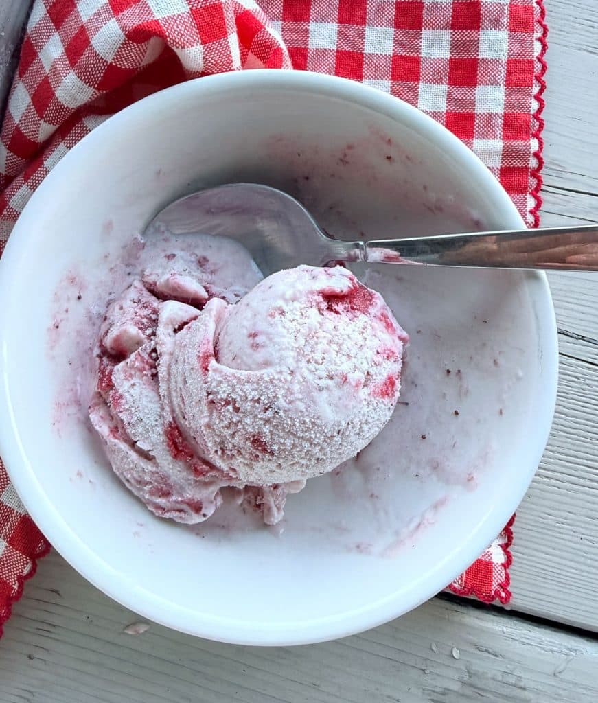 white bowl of strawberry ice cream startign to melt with a silver spoon in the bowl, a red and white gingham cloth underneath the bowl 