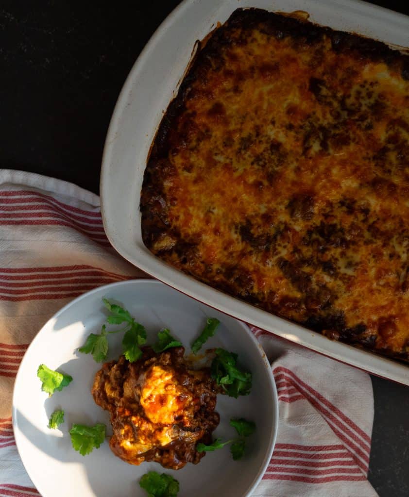 beef enchiladas in a casserole dish with a plate of enchiladas sitting next to the dish with a red and white striped towel under the dish on a black countertop