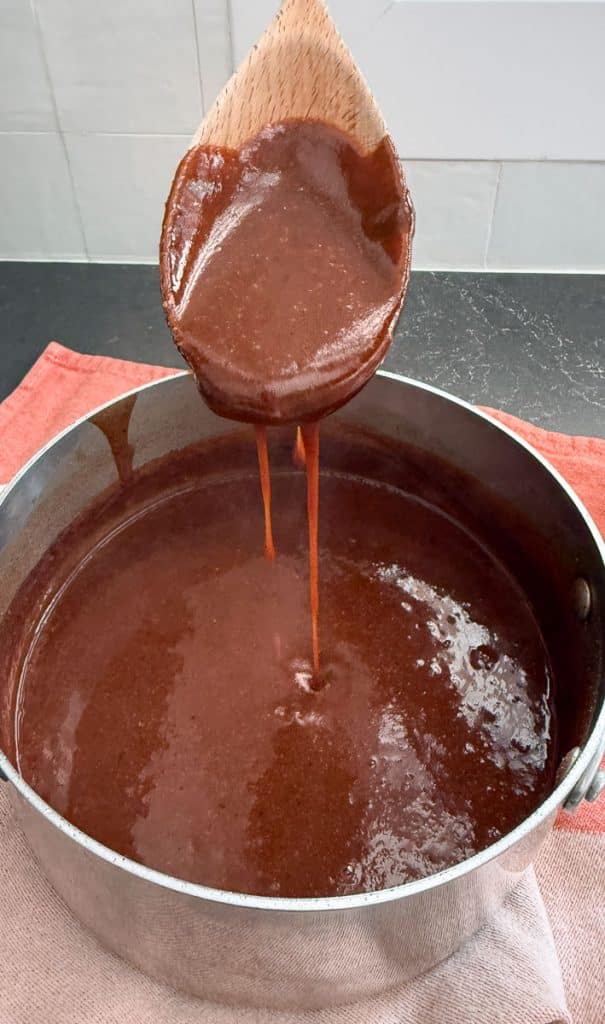 stirring enchilada sauce in pan there is a wood spoon hanging over the pan with sauce dripping off of it into the pan