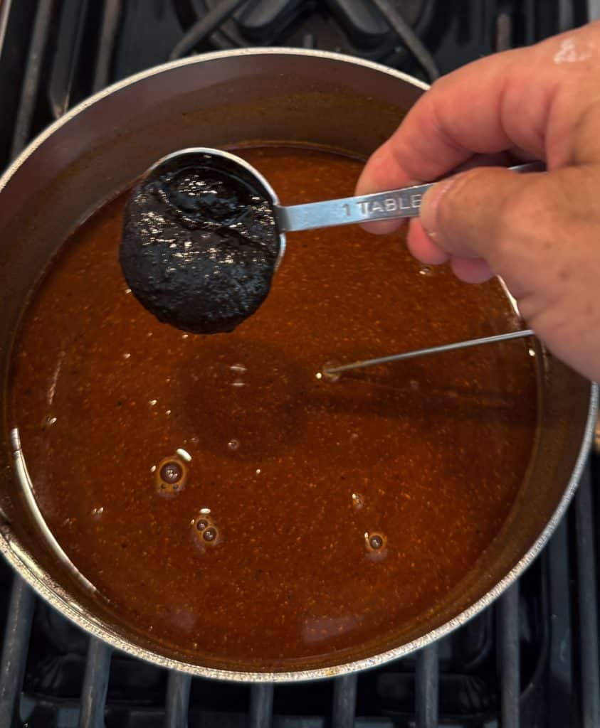 hand holding a tablespoon is adding beef bouillon to the red enchilada sauce