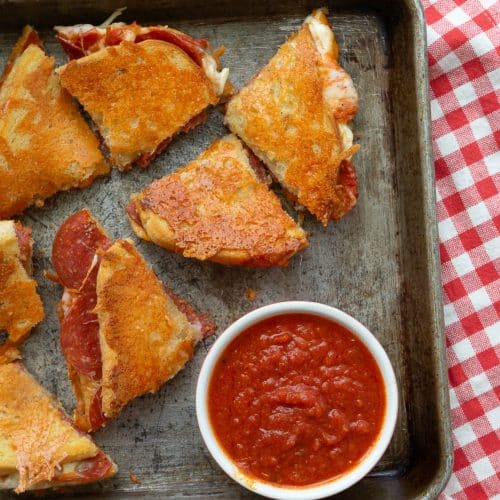 wedges of parmesan crusted pepperoni pizza grilled cheese on a baking sheet with a small bowl of pizza sauce in the lower right hand corner baking sheet is on a red and white gingham cloth