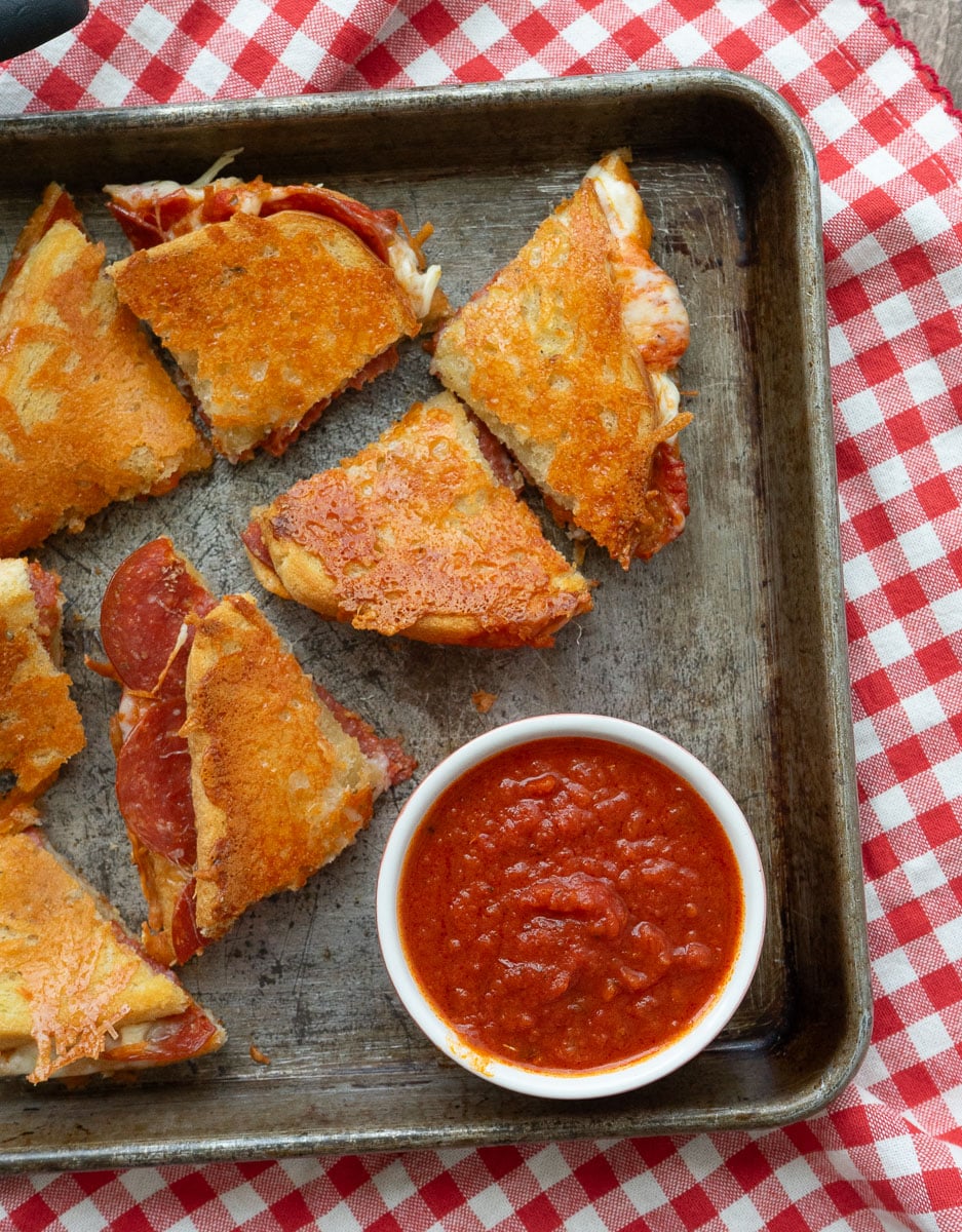 wedges of parmesan crusted pepperoni pizza grilled cheese on a baking sheet with a small bowl of pizza sauce in the lower right hand corner baking sheet is on a red and white gingham cloth