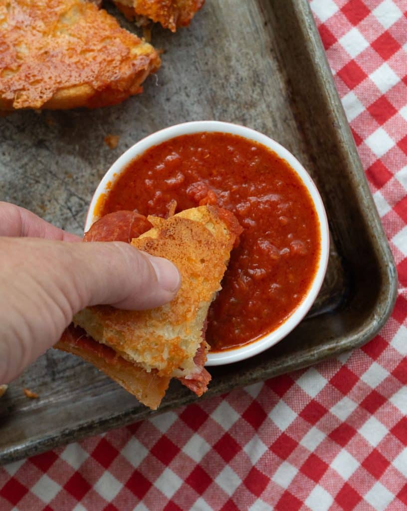 dipping grilled cheese into pizza sauce that is in a small dish sitting on a baking sheet full of grilled cheese wedges