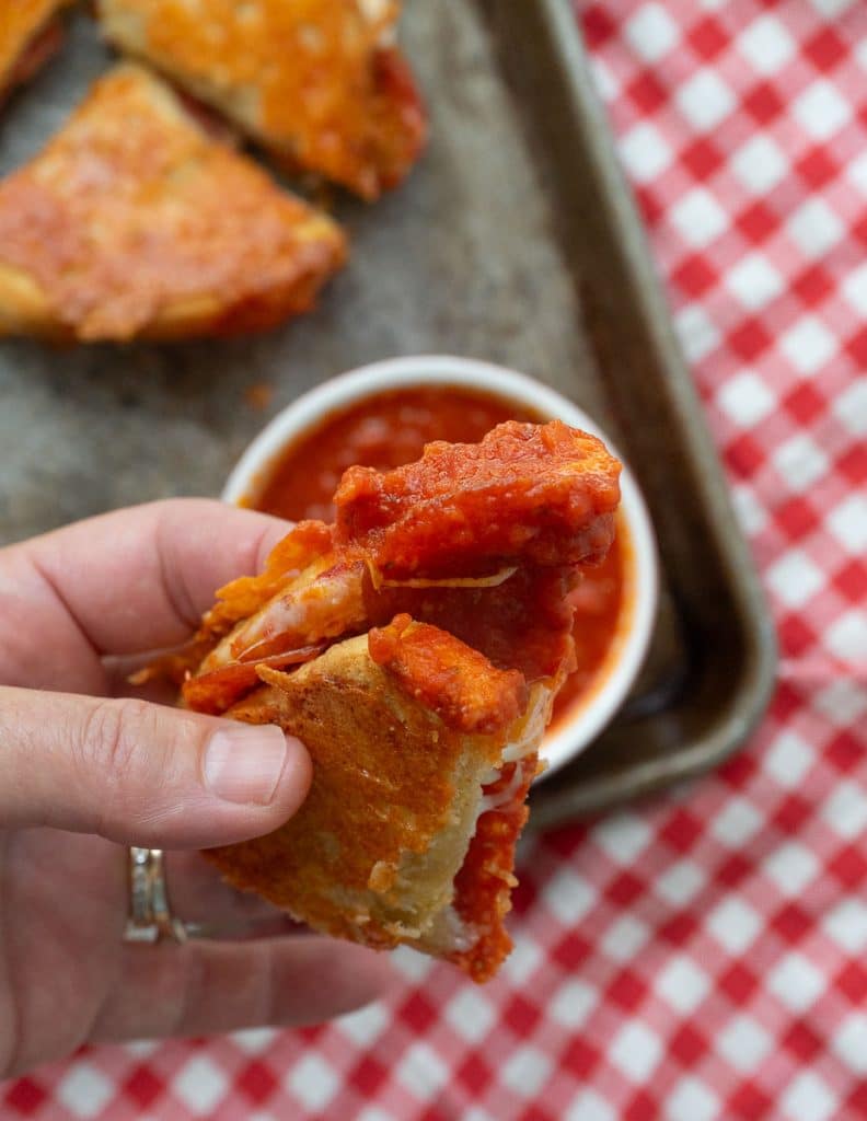 hand holding a wedge of grilled cheese that has been dipped in pizza sauce. Pan of grilled cheeses is in the background sitting on a red and white gingham cloth