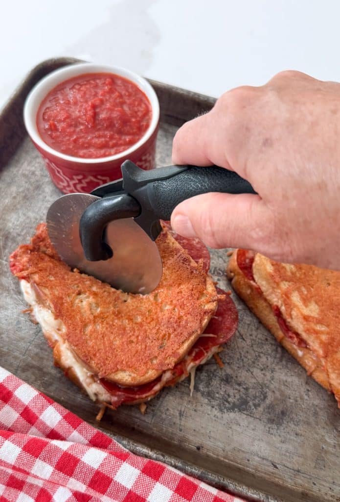 hand cutting sandwich with a pizza cutter on the baking sheet there is a small dish of pizza sauce in the upper left corner of the pan