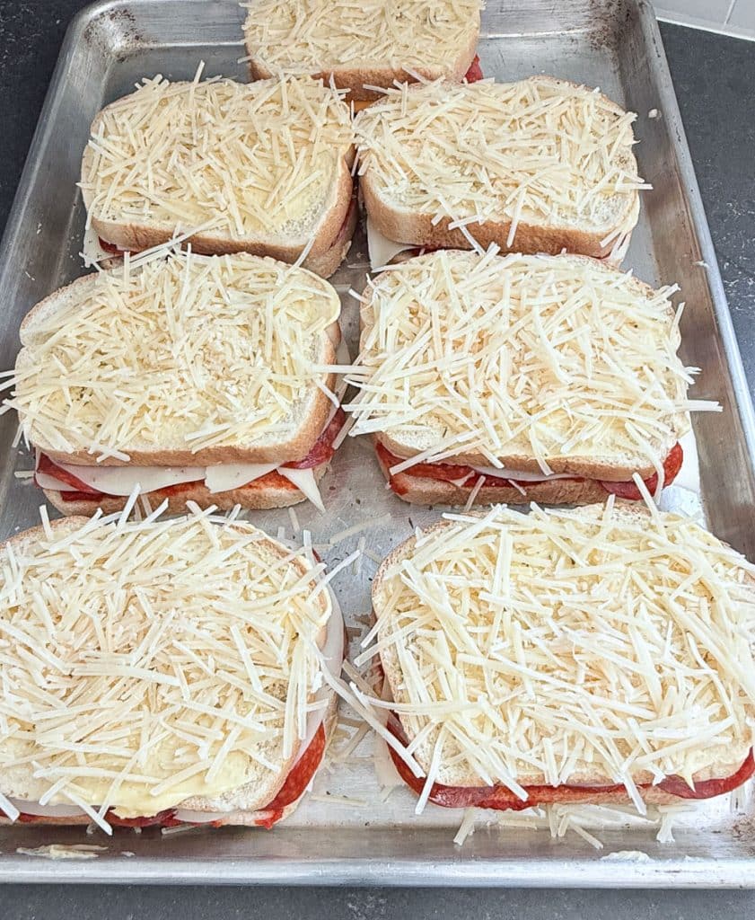 top slice of bread buttered and covered in shredded parmesan cheese is placed on top of the sandwich on a baking sheet