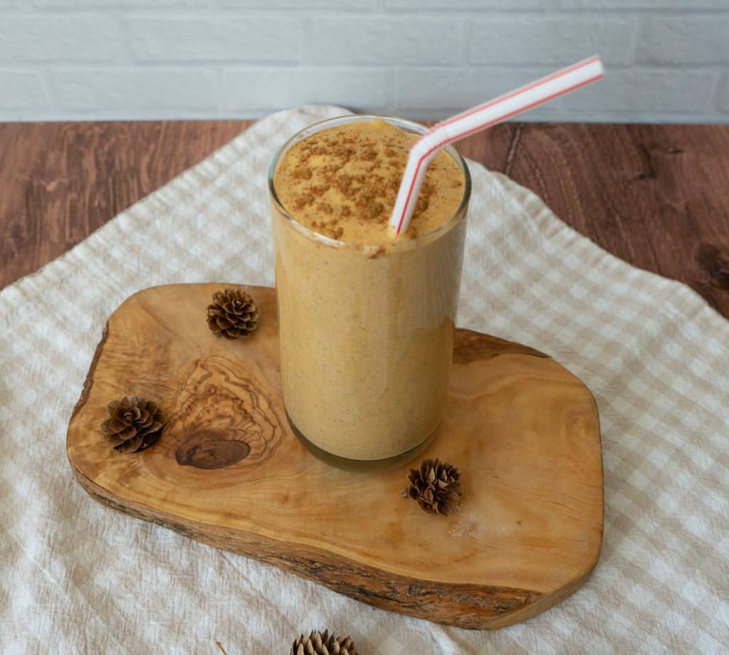 glass of pumpkin spice smoothie on a wood board with a straw sticking out to the right side the backsplash is a white tile and the counter top is wood, there is also a white and tan gingham cloth in the photo