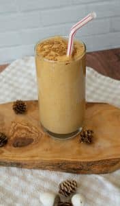 glass of pumpkin spice smoothie on a wood board with a straw sticking out to the right side the backsplash is a white tile and the counter top is wood, there is also a white and tan gingham cloth in the photo