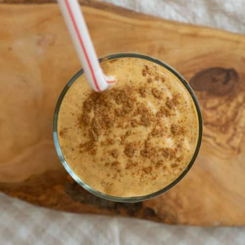 top view of a glass with pumpkin spice smoothie and a straw sticking out the glass is sitting on a wood board and there is a white and tan cloth underneath that