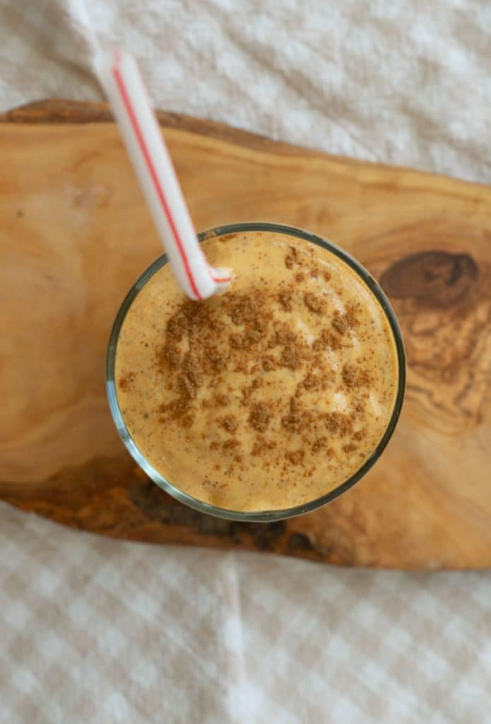 top view of a glass with pumpkin spice smoothie and a straw sticking out the glass is sitting on a wood board and there is a white and tan cloth underneath that