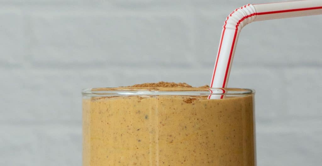 top of the glass that is full withe the pumpkin spice protein smoothie there is a straw sticking out of the top of the glass the back ground is a white tile