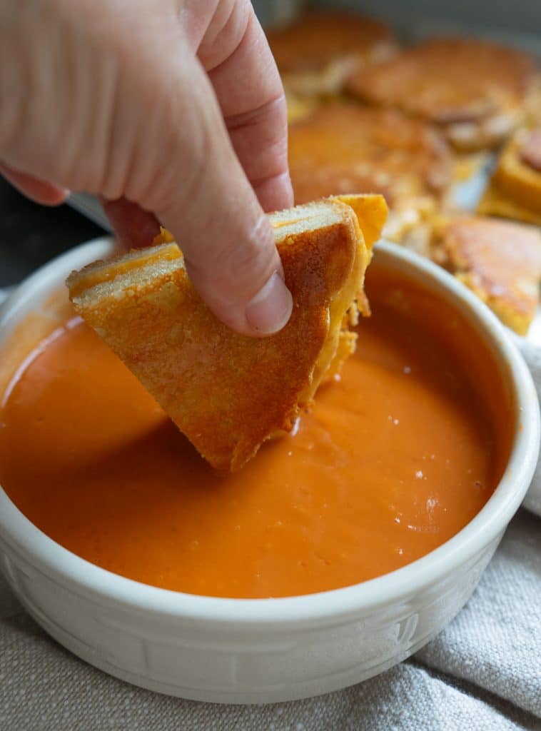 A hand holding a triangle of grilled cheese sandwich. dipping triangle in soup. there is a sheet pan of grilled cheese sandwiches in the back ground