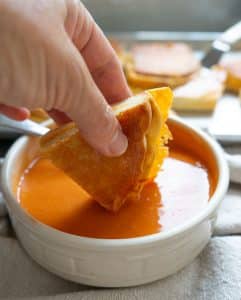hand holding a triangle of grilled cheese sandwich over a bowl of tomato soup, dipping triangle in soup. there is a sheet pan of grilled cheese sandwiches in the back ground