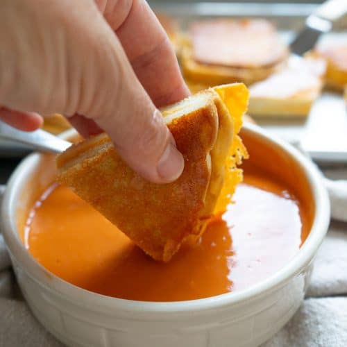 hand holding a triangle of grilled cheese sandwich over a bowl of tomato soup, dipping triangle in soup. there is a sheet pan of grilled cheese sandwiches in the back ground