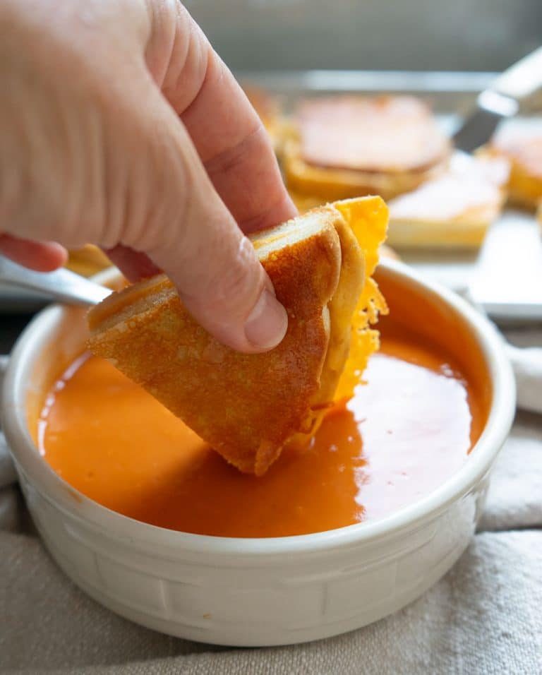 hand holding a triangle of grilled cheese sandwich over a bowl of tomato soup, dipping triangle in soup. there is a sheet pan of grilled cheese sandwiches in the back ground