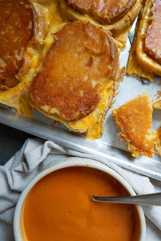 sheet pan of grilled cheese sandwiches next to a bowl of tomato soup