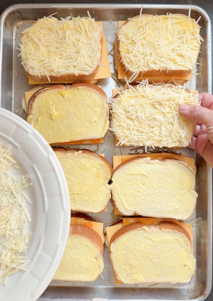 placing the top layer of parmesan crusted bread on to of grilled cheese sandwiches on baking sheet. hand is placing a piece of bread on top of another piece
